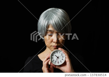 A senior woman with white hair, holding a round watch and looking down. A senior woman with white hair, holding a round watch and looking down. 137838514
