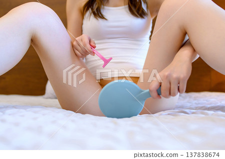 一位年輕女子在臥室裡對著手鏡,用 T 型剃刀刮比基尼部位的毛髮,進行自我照顧。 一位年輕女子在臥室裡對著手鏡,用 T 型剃刀刮比基尼部位的毛髮,進行自我照顧。 137838674