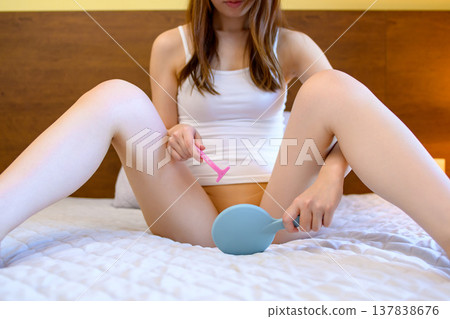 A young woman performs self-care on her VIO (bikini area) in her bedroom, shaving with a T-shaped razor while looking in a hand mirror. A young woman performs self-care on her VIO (bikini area) in her bedroom, shaving with a T-shaped razor while looking in a hand mirror. 137838676
