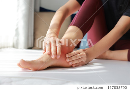 Woman sitting on floor and massaging her painful ankle with red highlight. Ankle sprain, joint inflammation, tendonitis and injury concept for health care, physical therapy and orthopedic treatment. Woman sitting on floor and massaging her painful ankle with red highlight. Ankle sprain, joint inflammation, tendonitis and injury concept for health care, physical therapy and orthopedic treatment. 137838790