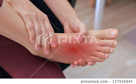 Woman sitting on floor and massaging her painful ankle with red highlight. Ankle sprain, joint inflammation, tendonitis and injury concept for health care, physical therapy and orthopedic treatment. Woman sitting on floor and massaging her painful ankle with red highlight. Ankle sprain, joint inflammation, tendonitis and injury concept for health care, physical therapy and orthopedic treatment. 137838845