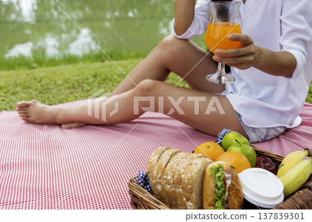 Close up Women picnic in green park hands holding glass of orange juice fresh fruit cool summer. Women drinking sit blanket with Picnic basket bread coffee cup in wood wicker healthy box red Blanket Close up Women picnic in green park hands holding glass of orange juice fresh fruit cool summer. Women drinking sit blanket with Picnic basket bread coffee cup in wood wicker healthy box red Blanket 137839301