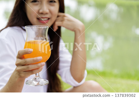 Women hands drink orange juice fresh fruit cool drinking in green park garden. Asian women hands holding healthy drinking glass. Beautiful female pouring orange juice from cocktail bottle outdoors Women hands drink orange juice fresh fruit cool drinking in green park garden. Asian women hands holding healthy drinking glass. Beautiful female pouring orange juice from cocktail bottle outdoors 137839302
