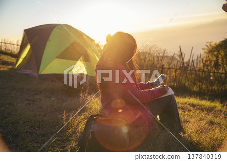 Women camping outdoor sitting Tent Camp trip hiking mountain hill on sunrise. Tent camp in nature background flare sunlight mountain. Picnic green nature campground Adventure Travel wildness forest Women camping outdoor sitting Tent Camp trip hiking mountain hill on sunrise. Tent camp in nature background flare sunlight mountain. Picnic green nature campground Adventure Travel wildness forest 137840319