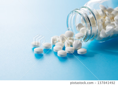 White tablets (supplements/medicine) spilling from a bottle placed against a blue background. White tablets (supplements/medicine) spilling from a bottle placed against a blue background. 137841492