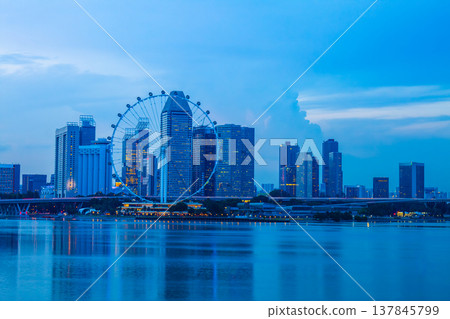 A modern building with a giant Ferris wheel by the sea, at twilight, overlooking the Singapore skyline. 137845799