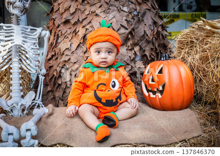 A little boy wearing a pumpkin costume and hat sits on a woven cloth to celebrate Halloween with a background of pumpkins, ghosts and skeletons. A little boy wearing a pumpkin costume and hat sits on a woven cloth to celebrate Halloween with a background of pumpkins, ghosts and skeletons. 137846370