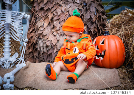 A little boy wearing a pumpkin costume and hat sits on a woven cloth to celebrate Halloween with a background of pumpkins, ghosts and skeletons. A little boy wearing a pumpkin costume and hat sits on a woven cloth to celebrate Halloween with a background of pumpkins, ghosts and skeletons. 137846384