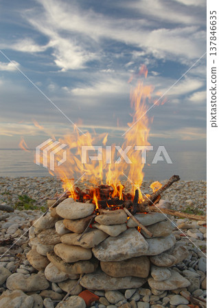 Bright campfire burning in stone ring on pebble beach at sunset with copy space Bright campfire burning in stone ring on pebble beach at sunset with copy space 137846635