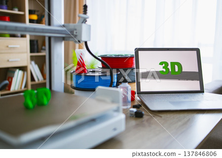 A 3D printer is actively creating a green object on its build plate next to spools of filament and a laptop displaying the 3D logo 137846806