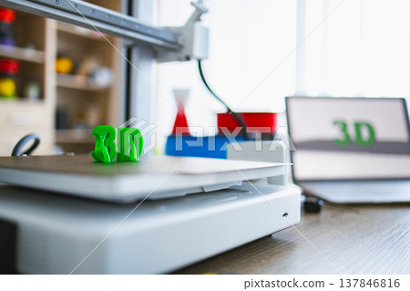 A 3D printer is actively creating a green plastic object on its build plate with a laptop displaying the 3D logo in the background A 3D printer is actively creating a green plastic object on its build plate with a laptop displaying the 3D logo in the background 137846816