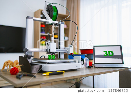 A modern 3D printer is actively creating green plastic letters on its build plate while a laptop displays the word 3D 137846833