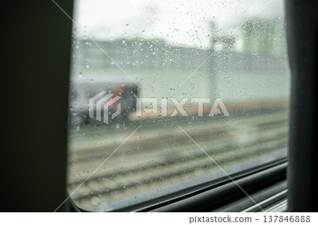 Raindrops cling to a train window while the vehicle passes through a city area, showcasing the blurred scenery outside on a grey day. Raindrops cling to a train window while the vehicle passes through a city area, showcasing the blurred scenery outside on a grey day. 137846888
