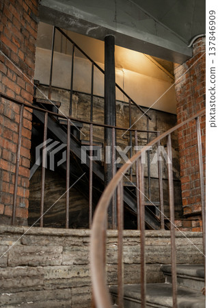 Sturdy spiral staircase ascends to a dimly lit upper level within an industrial setting, surrounded by exposed brick and rustic architecture. 137846909
