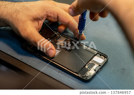 Technician carefully repairs smartphone by removing components with a screwdriver at a mobile repair workshop during daylight hours. Technician carefully repairs smartphone by removing components with a screwdriver at a mobile repair workshop during daylight hours. 137846957
