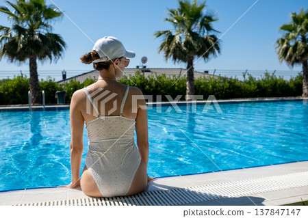 A woman enjoys a peaceful moment sitting by an outdoor pool, sunshine illuminating her and the surrounding palm trees creating a tropical vibe. A woman enjoys a peaceful moment sitting by an outdoor pool, sunshine illuminating her and the surrounding palm trees creating a tropical vibe. 137847147