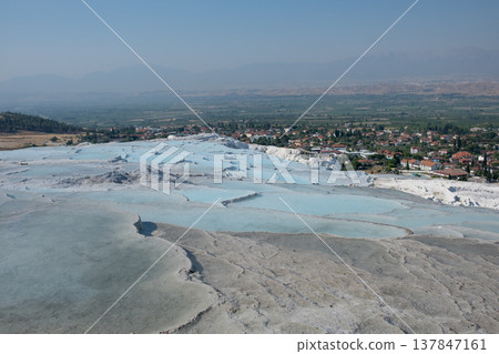 Pamukkale showcases its famous white travertine terraces and hot springs under a bright sky, with a picturesque village visible in the background. 137847161