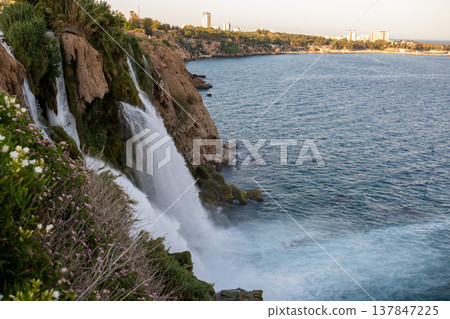 Waterfalls tumble down rocky cliffs into the turquoise Mediterranean Sea as the sun sets over Antalya, creating a picturesque coastal view filled with nature. 137847225