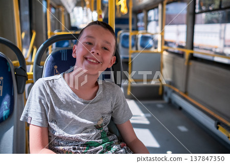 A cheerful young boy is sitting on a public bus, smiling joyfully. The interior features bright colors and spacious seating during the daytime. 137847350