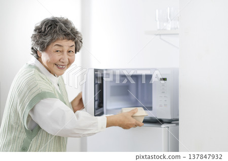 Senior woman heats food in microwave oven, looking directly at the camera. Senior woman heats food in microwave oven, looking directly at the camera. 137847932