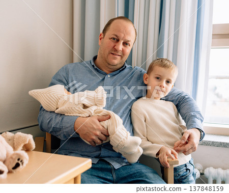 Father sitting with older son while holding newborn baby girl in hospital room. Warm family moment showing love and connection between siblings and parents. Father sitting with older son while holding newborn baby girl in hospital room. Warm family moment showing love and connection between siblings and parents. 137848159