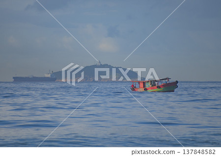 Traditional Colorful Fishing Boat in the Sea with Tropical Island and Cargo Ship on Horizon Traditional Colorful Fishing Boat in the Sea with Tropical Island and Cargo Ship on Horizon 137848582