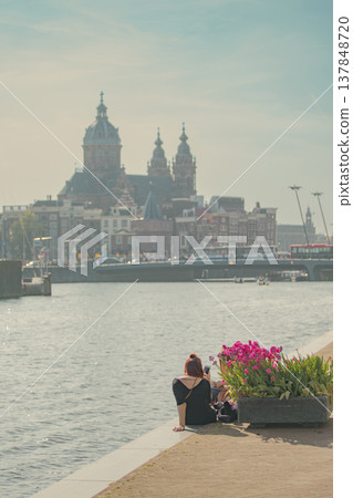Young woman traveler sitting by canal admiring Basilica of Saint Nicholas and tulips in Amsterdam with copy space 137848720