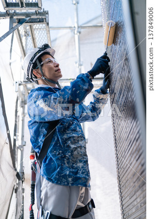 A male craftsman works at a site painting an exterior wall. He is diligently repainting, his work clothes covered in paint. A male craftsman works at a site painting an exterior wall. He is diligently repainting, his work clothes covered in paint. 137849006