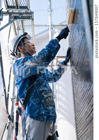 A male craftsman works at a site painting an exterior wall. He is diligently repainting, his work clothes covered in paint. A male craftsman works at a site painting an exterior wall. He is diligently repainting, his work clothes covered in paint. 137849007