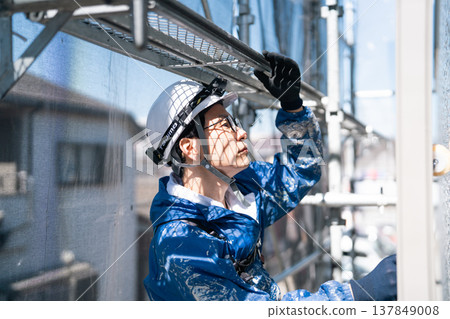 A male craftsman works at a site painting an exterior wall. He is diligently repainting, his work clothes covered in paint. 137849008