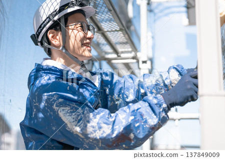 A male craftsman works at a site painting an exterior wall. He is diligently repainting, his work clothes covered in paint. 137849009