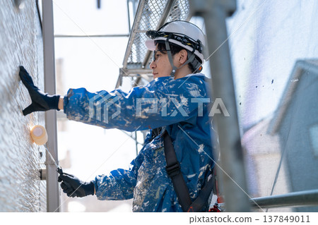 A male craftsman works at a site painting an exterior wall. He is diligently repainting, his work clothes covered in paint. A male craftsman works at a site painting an exterior wall. He is diligently repainting, his work clothes covered in paint. 137849011