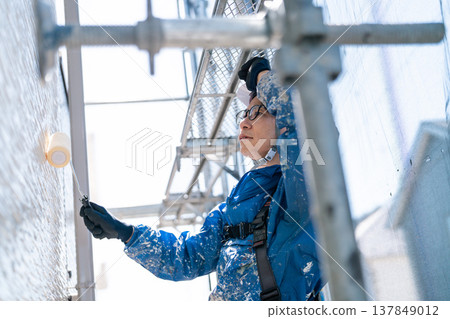 A male craftsman works at a site painting an exterior wall. He is diligently repainting, his work clothes covered in paint. 137849012