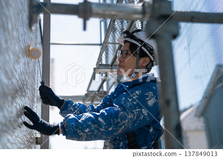 A male craftsman works at a site painting an exterior wall. He is diligently repainting, his work clothes covered in paint. 137849013
