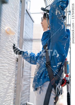 A male craftsman works at a site painting an exterior wall. He is diligently repainting, his work clothes covered in paint. A male craftsman works at a site painting an exterior wall. He is diligently repainting, his work clothes covered in paint. 137849016