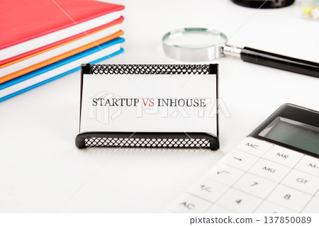 White desk holds STARTUP VS INHOUSE card, calculator, magnifying glass, colorful notebooks. Strategic business choices White desk holds STARTUP VS INHOUSE card, calculator, magnifying glass, colorful notebooks. Strategic business choices 137850089