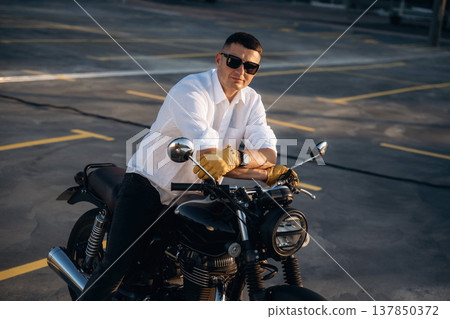 White shirt, black sunglasses. Man is outdoors with his motorcycle White shirt, black sunglasses. Man is outdoors with his motorcycle 137850372