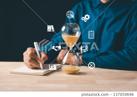 Hourglass shows time management concept, person writes notes on book, planning for business deadline countdown, sand flowing through glass, managing important task efficiently Hourglass shows time management concept, person writes notes on book, planning for business deadline countdown, sand flowing through glass, managing important task efficiently 137850696