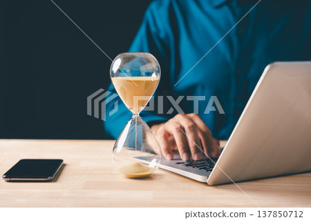 Hourglass shows time passing, sand flowing, urgency for business growth, deadline countdown drives person typing on laptop, optimizing work for success, future strategic planning 137850712