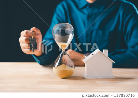 Hand holds real estate house keys, hourglass with falling sand marks time, symbolizing mortgage deadline, home buying, property loan concept for urgent housing investment 137850716