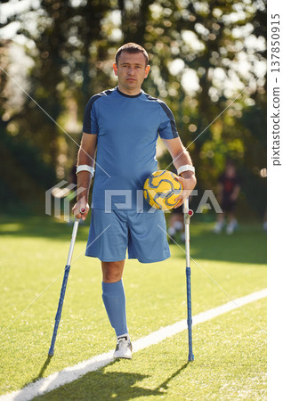 Holding soccer ball. Disabled man with crutches is on the field Holding soccer ball. Disabled man with crutches is on the field 137850915