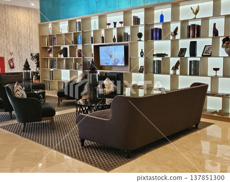 A sophisticated hotel lounge featuring a large illuminated bookshelf wall, plush dark velvet sofas, and elegant decor items, creating a warm and luxurious atmosphere for guests in a modern interior. 137851300