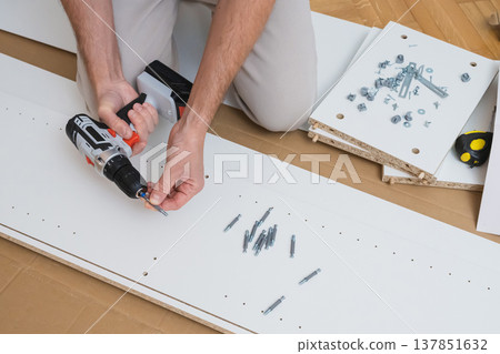 Male hands use a screwdriver to fasten hardware into a white furniture panel with assembly parts and instructions laid out on a wooden floor Male hands use a screwdriver to fasten hardware into a white furniture panel with assembly parts and instructions laid out on a wooden floor 137851632