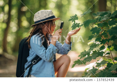Discovering plants under magnifying glass. Young girl hiker is in the forest Discovering plants under magnifying glass. Young girl hiker is in the forest 137852439