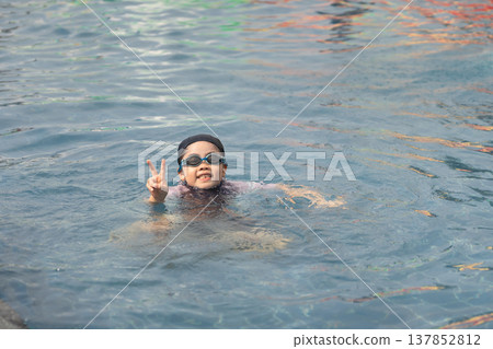 Young child enjoying swimming in a vibrant pool, making a cheerful peace sign with fingers while wearing goggles, capturing a moment of joy and fun Young child enjoying swimming in a vibrant pool, making a cheerful peace sign with fingers while wearing goggles, capturing a moment of joy and fun 137852812