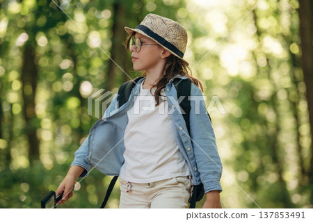 In hat and with backpack. Young girl hiker is in the forest In hat and with backpack. Young girl hiker is in the forest 137853491