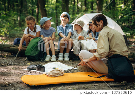 Book in hand, sitting together. Woman is doing tour for group of kids in the forest Book in hand, sitting together. Woman is doing tour for group of kids in the forest 137853610
