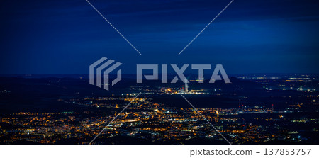 Aerial view of a city illuminated at night with glowing lights, distant mountains, and a power plant emitting steam in the background under a twilight sky Aerial view of a city illuminated at night with glowing lights, distant mountains, and a power plant emitting steam in the background under a twilight sky 137853757