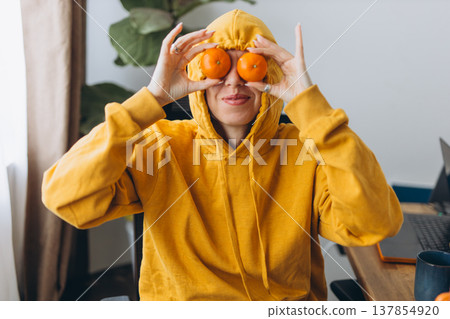 Creative woman with tangerines over eyes in hoodie showing playful viral content idea in home office with modern lifestyle vibe 137854920