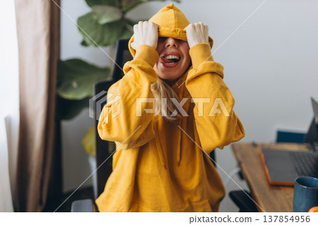 Playful woman covering face with hoodie making funny expression in home office showing stress relief and carefree lifestyle moment Playful woman covering face with hoodie making funny expression in home office showing stress relief and carefree lifestyle moment 137854956
